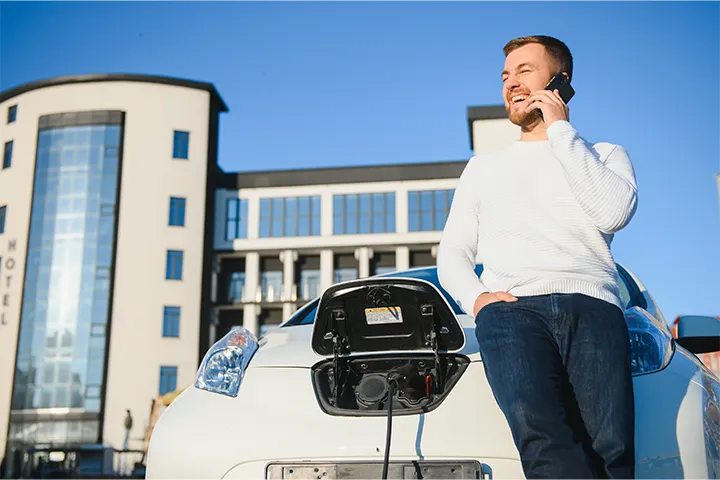 Handsome person talking with someone via phone while his ev is charging by Xova Chargers in front of multifamily complex